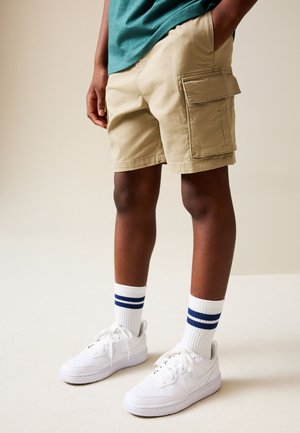 Person iført beige cargoshorts, hvide sneakers og hvide crew-sokker med to blå striber, står mod en ensfarvet baggrund.