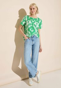 Camicia a maniche corte verde e bianca a fantasia, abbinata a jeans a gamba larga blu chiaro e sneakers bianche, su uno sfondo beige.