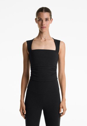 Black fitted tank top with square neckline and wide straps, featuring gathered texture at the waist. Worn with matching black high-waisted leggings.