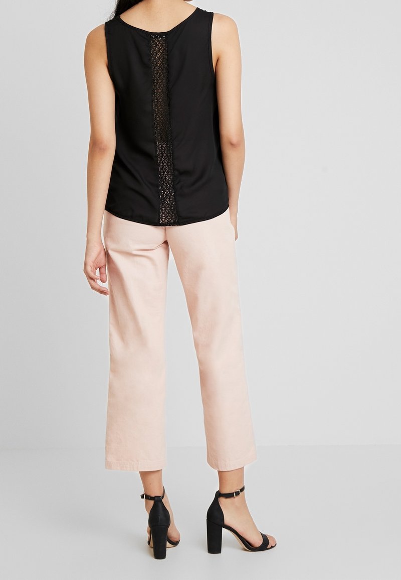 Černý tílko bez rukávů s krajkovým detailem na zádech, kombinované s lehkými růžovými kalhotami širokého střihu a černými sandály na podpatku. Minimalistický design.