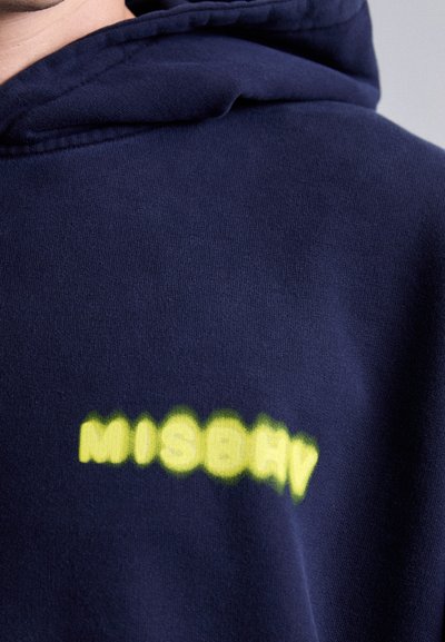 Námornícky modrá mikina s kapucňou so žltým rozmazaným textom "MISBHV" na hrudi, zobrazuje štruktúru látky a časť kapucne pri krku.