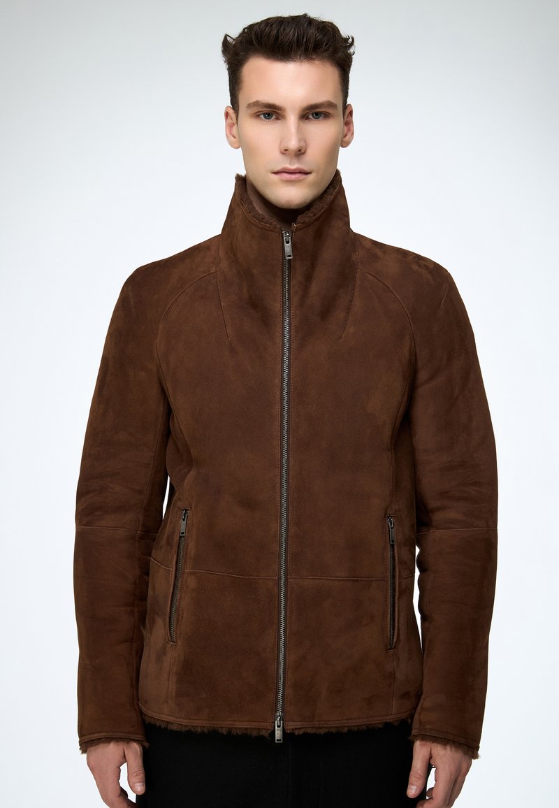 Junger Mann, der eine braune Wildlederjacke mit Reißverschluss und hohem Kragen trägt, vor einem schlichten hellen Hintergrund stehend.