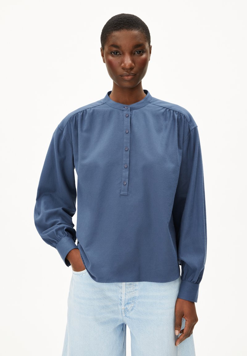 Donna che indossa una camicia blu a maniche lunghe con maniche a sbuffo e jeans azzurri, in piedi con una mano in tasca.