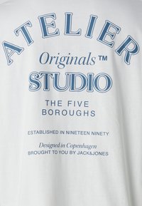 Λευκή βαμβακερή μπλούζα με μπλε κείμενο. Στο πίσω μέρος αναγράφεται "ATELIER Originals STUDIO THE FIVE BOROUGHS" και άλλες λεπτομέρειες σε έντονη γραφή.