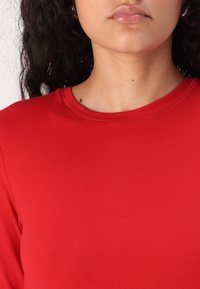 Bluză roșie cu mâneci lungi, confecționată dintr-un material neted și elastic, cu decolteu rotund și detalii de cusătură subtile, care se îmbină strâns cu corpul.