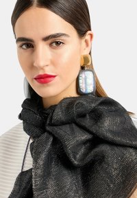 Sciarpa nera con una finitura metallica texturizzata, abbinata a orecchini oversize dorati e iridescenti. Pelle con toni naturali e labbra rosse.
