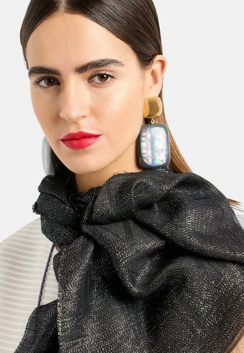 Sciarpa nera con una finitura metallica texturizzata, abbinata a orecchini oversize dorati e iridescenti. Pelle con toni naturali e labbra rosse.