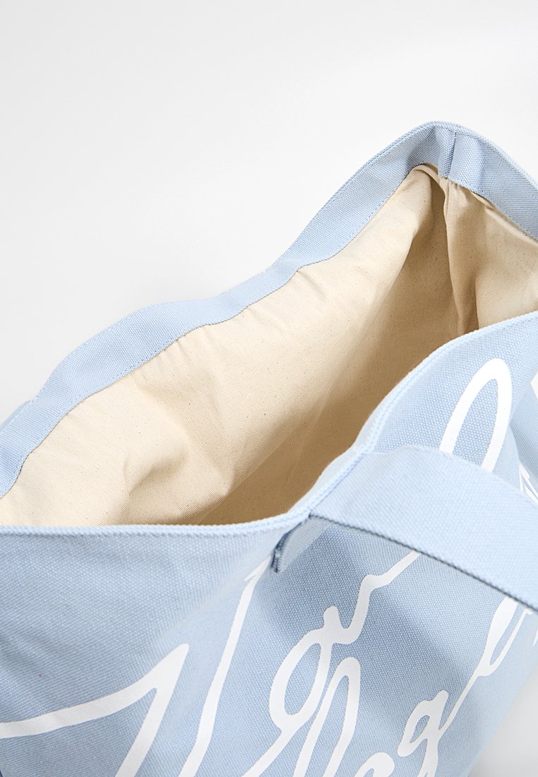 Hellblaues Canvas-Shopper mit beigem Innenfutter, weißer geschwungener Schrift auf der Außenseite und stabilen Griffen vor einem weißen Hintergrund.