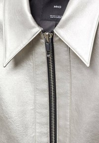 Veste en cuir argentée avec col pointu et fermeture éclair centrale argentée, montrant la doublure intérieure noire et l'étiquette de la marque MNG.