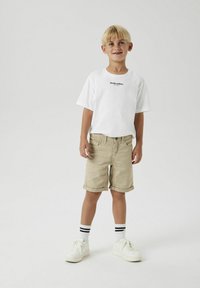 T-shirt blanche à manches courtes avec un imprimé textuel noir, shorts beige à revers, baskets blanches et chaussettes à rayures noires et blanches.