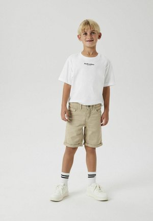 T-shirt blanche à manches courtes avec un imprimé textuel noir, shorts beige à revers, baskets blanches et chaussettes à rayures noires et blanches.