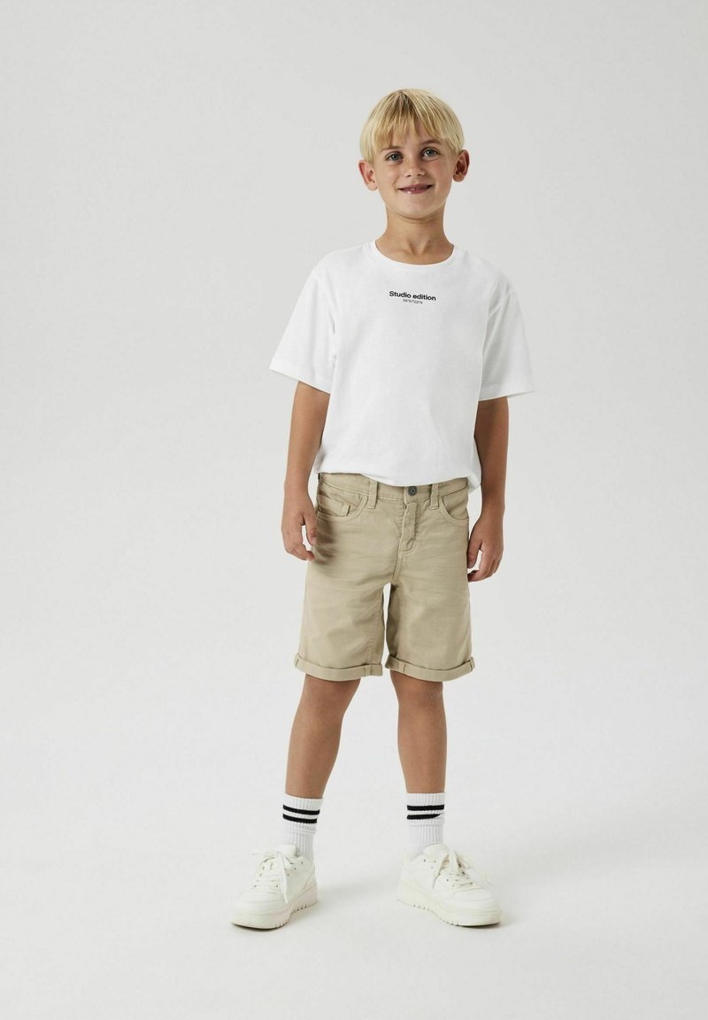 T-shirt blanche à manches courtes avec un imprimé textuel noir, shorts beige à revers, baskets blanches et chaussettes à rayures noires et blanches.