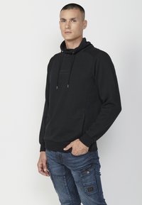 Sudadera negra con un patrón de espiga texturizado, cordón ajustable, bolsillo frontal y puños de canalé, combinada con pantalones cargo azules.