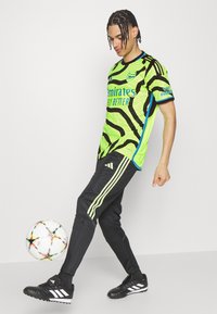 Camiseta de fútbol de manga corta con patrón verde y negro, acompañada de pantalones negros con rayas verdes y botas negras, junto a un balón de fútbol.