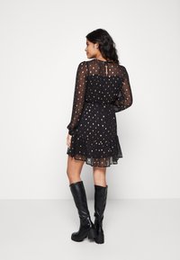 Robe noire transparente avec des pois dorés, ourlet volanté et manches longues. Fermeture en forme de clé au dos. Portée avec des bottes noires Haute jusqu'aux genoux.