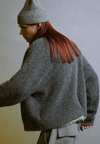 Maglione grigio in lana oversize con una finitura testurizzata, abbinato a pantaloni grigi. Un berretto a coste grigio completa l'outfit.