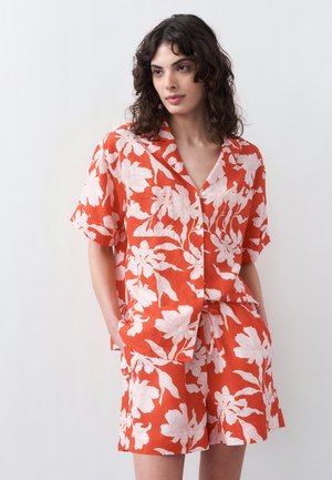 Kobieta o kręconych ciemnych włosach, ubrana w pomarańczową koszulę w kwiaty z krótkim rękawem na guziki oraz dopasowane spodenki, stojąca na jednolitym tle.