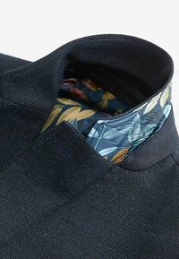 Collo in tessuto blu scuro con un rivestimento a fiori che presenta foglie nei toni del verde, marrone e oro. La superficie strutturata aggiunge profondità.