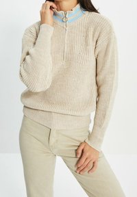 Beige gestrickter Pullover mit einem halben Reißverschlusskragen, der hellblaue Akzente aufweist. Die Textur ist grob, mit gerippten Bündchen und Saum.