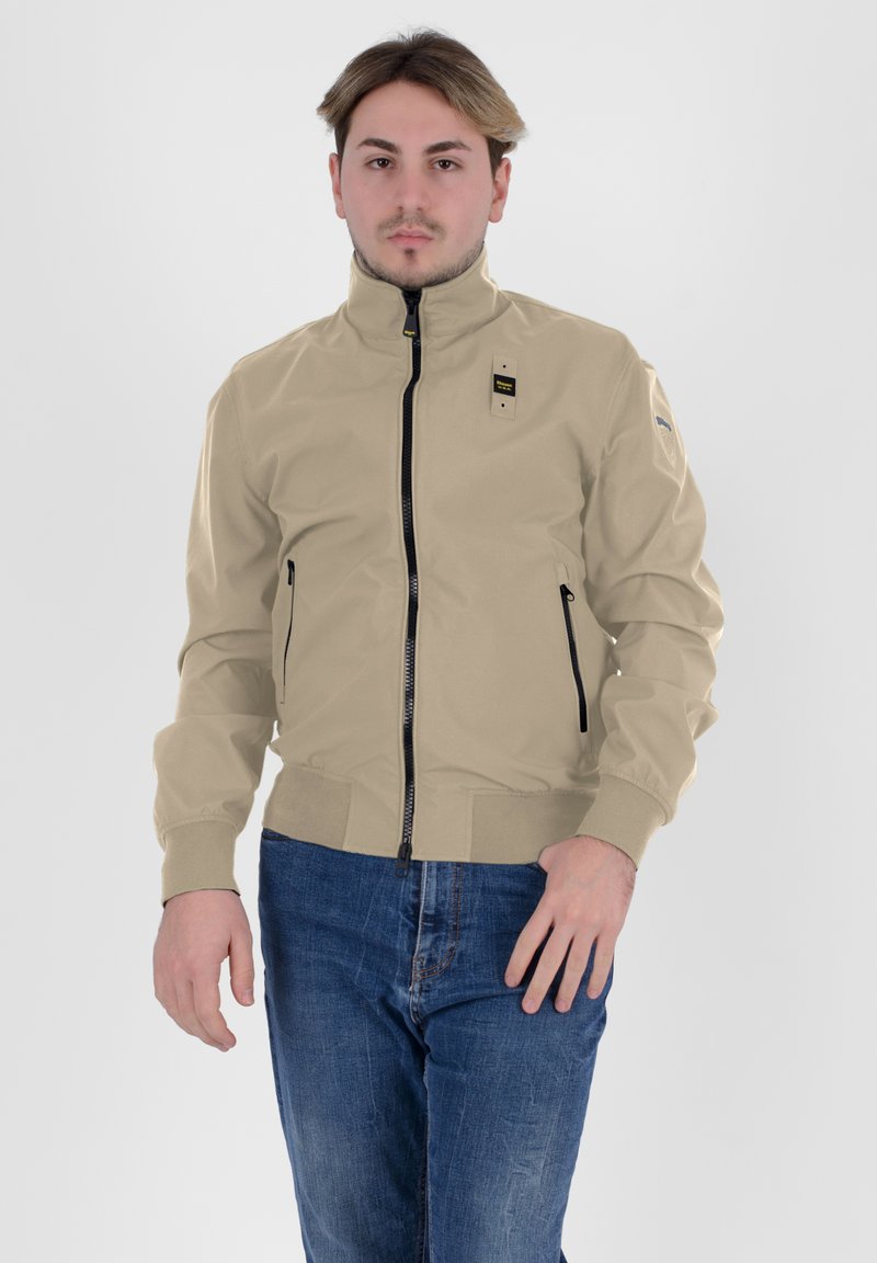 Giacca beige con colletto alto, zip frontale e tasche con zip; presenta accenti neri e un'etichetta sul petto; texture liscia.
