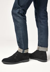 Black suede ankle boots with a flat sole and minimalist design, paired with dark denim jeans featuring rolled cuffs and contrasting stitching.