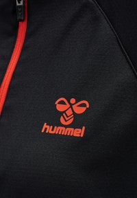 Giacca sportiva nera con zip rossa e un evidente logo Hummel arancione con design a farfalla e dettagli a chevron. Tessuto morbido.