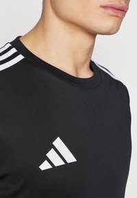 Zwart sport t-shirt met een ronde hals, drie witte strepen op de schouders en een wit Adidas-logo op de voorkant. Glad textuur.