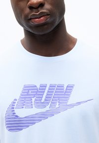 Κοντινή λήψη ενός ατόμου που φοράει λευκή μπλούζα με μοβ κείμενο "RUN" και λογότυπο swoosh στο στήθος.