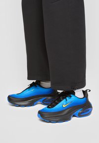 Scarpe sportive caratterizzate da un design blu e nero con una texture liscia, evidenziate da un logo Nike giallo e una suola arrotondata con ammortizzazione ad aria.
