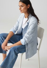 Blazer su misura azzurro chiaro con maniche arrotolate, indossato sopra una camicia bianca. Abbinato a jeans in denim blu larghi. Seduto su una sedia bianca.