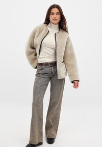 Bežs mākslīgā kažoks ar melniem akcentiem, brīva piegriezuma, ar nolaistām plecu šuvēm, uzvilkts virs bežā turtleneck un kombinēts ar gaiši brūnām platām džinsēm.