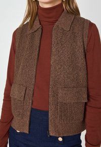 Gilet lavorato a maglia marrone con una texture a coste, design con colletto e due tasche frontali, abbinato a un dolcevita bordeaux e pantaloni blu navy.