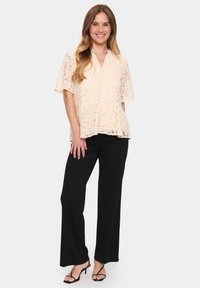 Blusa in pizzo color pesca chiaro con maniche corte e scollatura a V. Abbinata a pantaloni neri a gamba larga e sandali con tacco a strappo.