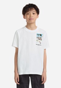 Baltas kokvilnas T-krekls ar priekšējo krūšu grafiku, kurā attēloti palmu koki, pludmales ainava un uzraksts "Abercrombie & Fitch". Īsas piedurknes, apaļš kakls.