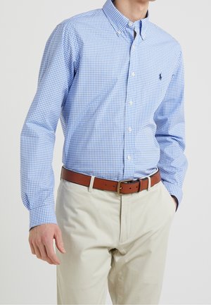 Homme portant une chemise à manches longues à carreaux bleu clair rentrée dans un pantalon beige avec une ceinture en cuir marron, debout avec une main dans la poche.