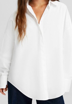 Witte langslavige button-up shirt met een spitse kraag, recht gesneden, oversized pasvorm en een zacht gestructureerde stof. De randen zijn afgerond.