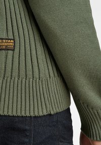 Olivgrüner Pullover mit gerippter Struktur, engem Saum und einem gewebten Etikett mit Markendetails an der Seite.