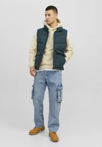 Padded dark teal vest over a beige hoodie, paired with light blue cargo jeans and tan boots. Features two front pockets and a zip closure.