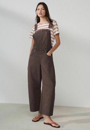 Junge Frau steht, trägt braune weite Latzhosen, ein gestreiftes Kurzarmshirt und orangefarbene Flip-Flops vor einem neutralen Hintergrund.