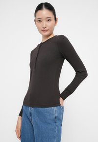 Jonge vrouw in een zwart langemouwen henley-shirt en blauwe jeans, staand met één hand in de zak tegen een witte achtergrond.
