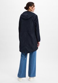 Navyblauwe waterdichte jas met een capuchon, een taille met aansnoerkoord, elastische manchetten en een langere achterkant. G gedragen met een wijde spijkerbroek en sneakers.