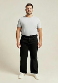 Bărbat stând cu fața înainte, purtând un tricou gri deschis, pantaloni negri și pantofi albi pe un fundal bej simplu.