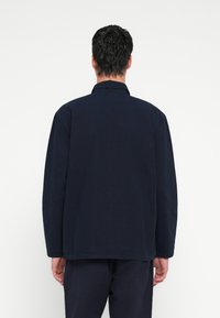 Μπλε ναυτικού τζάκετ με μακριά μανίκια και γιακά, κατασκευασμένο από απαλό ύφασμα. Ευθεία σιλουέτα με άνετη εφαρμογή και χωρίς ορατά διακοσμητικά στοιχεία.
