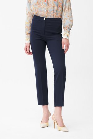 Kvinde iført skræddersyede navy ankel-lange bukser, en lyseblå mønstret bluse og beige høje hæle slingback-sko, stående mod en hvid baggrund.