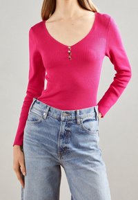 Frau in einem enganliegenden, langärmligen, gerippten rosa Oberteil mit drei dekorativen Knöpfen und hochgeschnittenen hellblauen Jeans vor einem grauen Hintergrund.