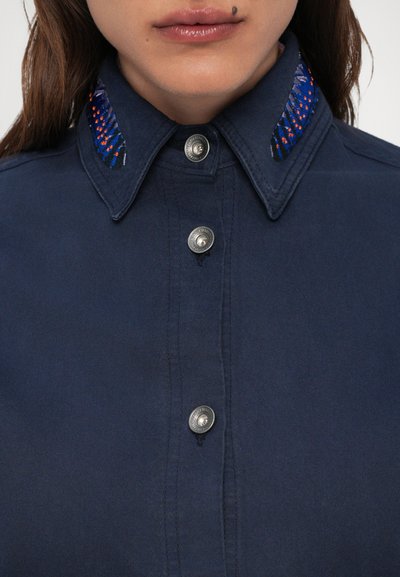 Κοντινό πλάνο από κόψιμο ενός ναυτικού πουκάμισου με ασημένια κουμπιά και κεντημένα μπλε και πορτοκαλί μοτίβα σε κάθε άκρη του γιακά.