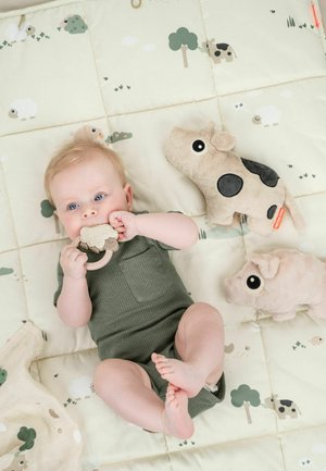 Un drap de lit vert clair pour lit d'enfant présente des moutons et des arbres dessinés en cartoon. Un bébé portant une grenouillère verte côtelée joue avec une tétine en forme de mouton beige et deux peluches.