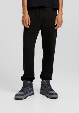 Homme portant un pantalon de jogging noir avec des poches avant et des baskets montantes gris foncé, debout sur un fond gris clair uni.