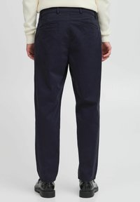 Navy blue chinos with a smooth texture, two back pockets, and a tailored fit, paired with black shoes, viewed from the back.