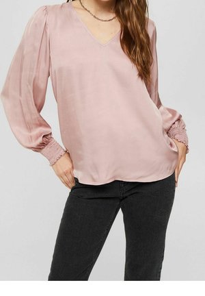 Frau trägt eine hellrosa V-Ausschnitt-Bluse mit langen, aufgepufften Ärmeln und gerafften Bündchen, kombiniert mit schwarzen Hosen vor einem schlichten Hintergrund.